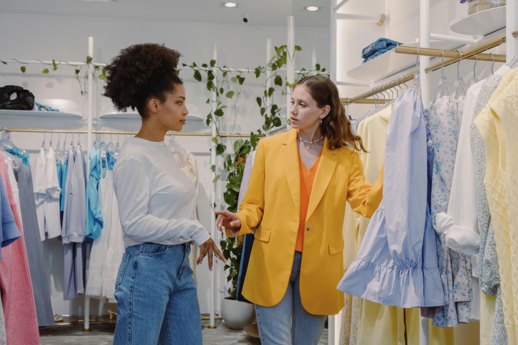 Two women in a boutique store browsing clothes, discussing styles.